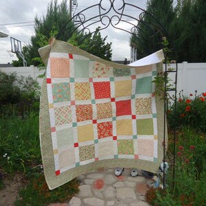 May include: A patchwork quilt featuring a grid of square fabric blocks in shades of peach, sage green, yellow, and red floral patterns. The quilt is bordered by a textured olive green fabric and accented with small red square cornerstones.