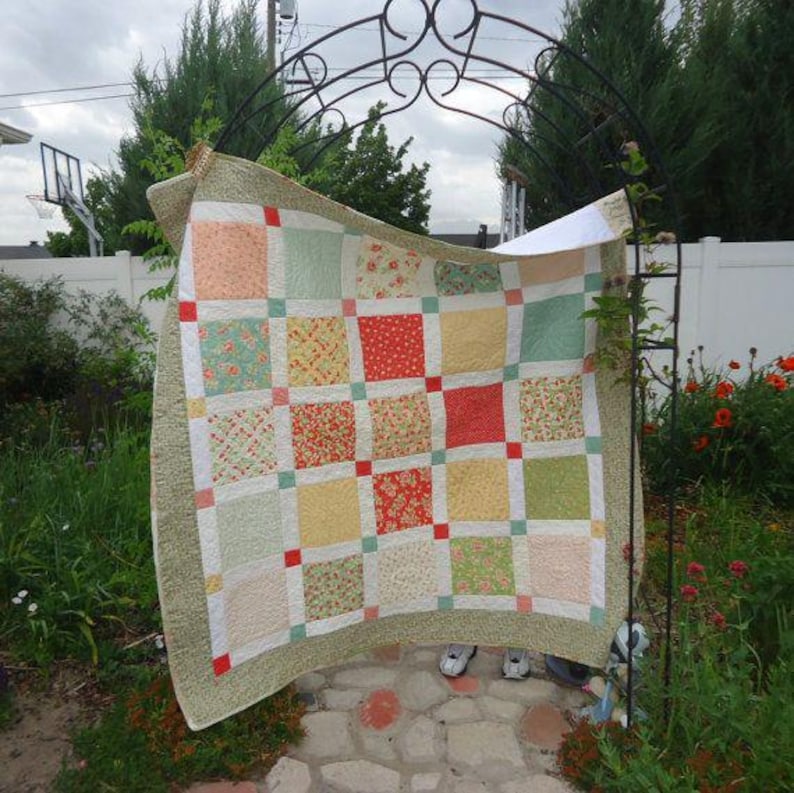 May include: A patchwork quilt featuring a grid of square fabric blocks in shades of peach, sage green, yellow, and red floral patterns. The quilt is bordered by a textured olive green fabric and accented with small red square cornerstones.