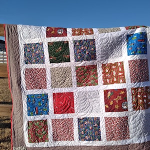 May include: A patchwork quilt featuring a grid of square fabric blocks with various Christmas-themed prints in red, green, blue, and white. The quilt is finished with a brown and white checkered border and white sashing with swirling stitching.