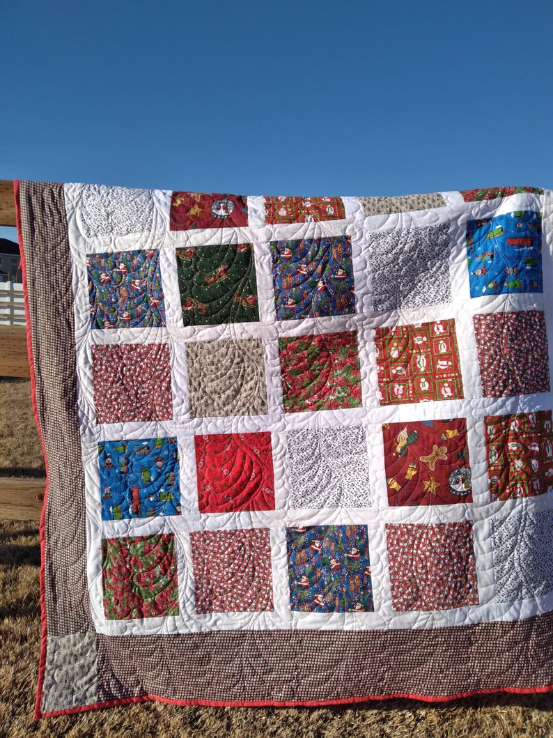 May include: A patchwork quilt featuring a grid of square fabric blocks with various Christmas-themed prints in red, green, blue, and white. The quilt is finished with a brown and white checkered border and white sashing with swirling stitching.