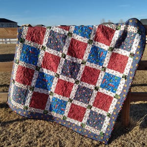 May include: A patchwork quilt featuring a grid of alternating solid red squares and blue squares patterned with small holiday-themed illustrations. The squares are separated by white sashing with a festive print and a dark blue star-patterned border.