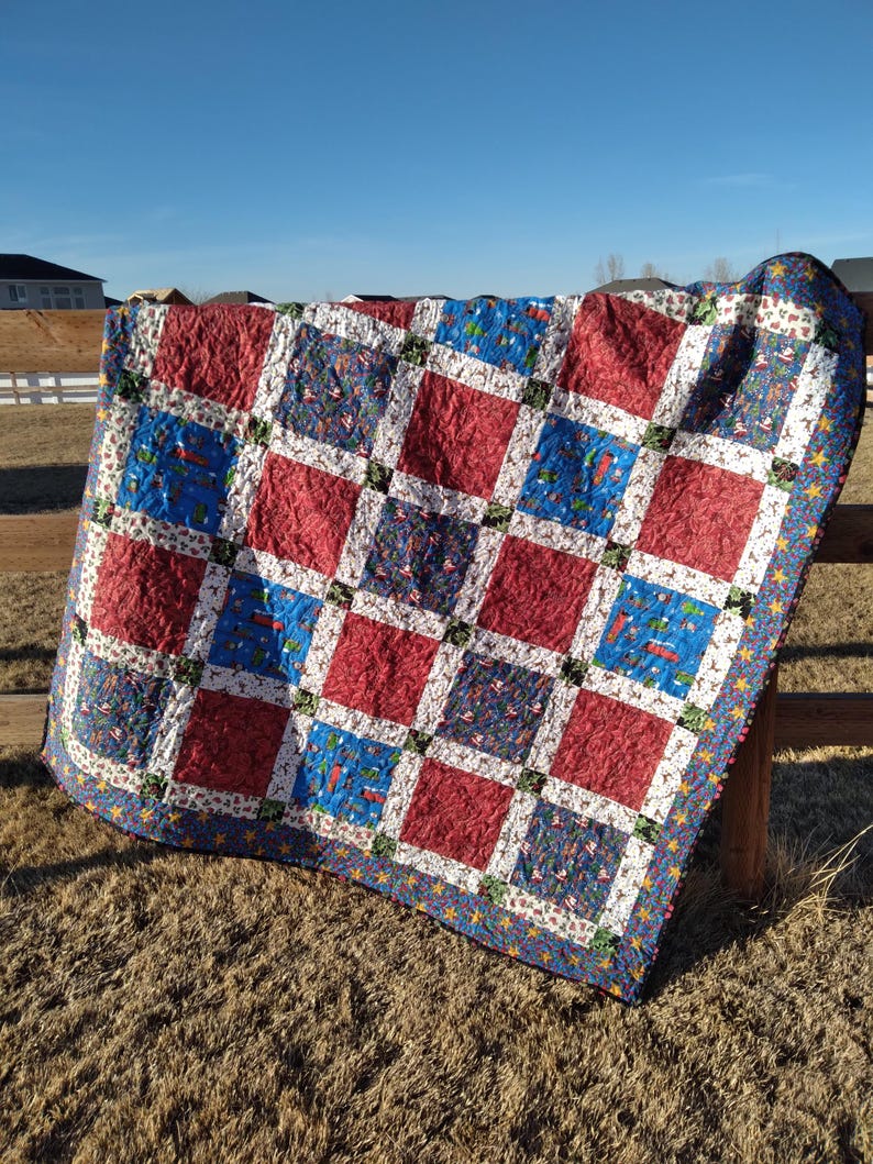 May include: A patchwork quilt featuring a grid of alternating solid red squares and blue squares patterned with small holiday-themed illustrations. The squares are separated by white sashing with a festive print and a dark blue star-patterned border.