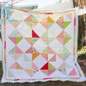 May include: A white quilt with a patchwork design featuring pink, green, red, and orange triangles. The quilt is edged with a green and white border. The quilt is hanging on a clothesline outside. The text "Modern Tradition Quilts" is visible in the top left corner of the quilt.