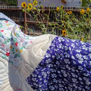 May include: A quilted fabric piece featuring a patchwork design with sections of white fabric printed with small red, yellow, and green floral patterns, solid cream-colored fabric with swirling stitched textures, and deep blue fabric with white floral prints.