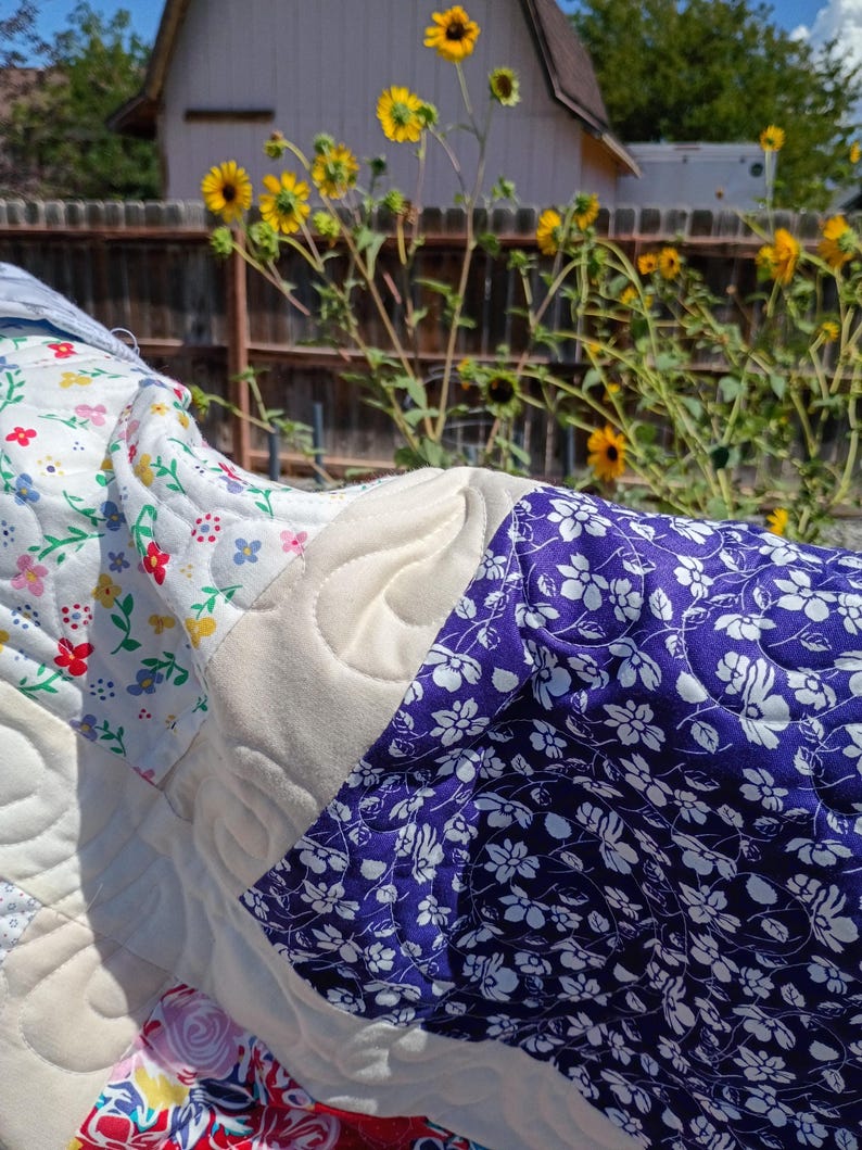 May include: A quilted fabric piece featuring a patchwork design with sections of white fabric printed with small red, yellow, and green floral patterns, solid cream-colored fabric with swirling stitched textures, and deep blue fabric with white floral prints.
