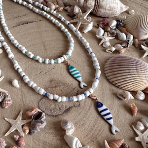 May include: Two white beaded necklaces with fish pendants. One pendant is turquoise with white stripes, the other is white with blue stripes. The necklaces are displayed with seashells and starfish on a wooden surface.