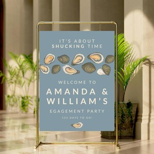 May include: A gold metal framed sign with a blue background. The sign reads "IT'S ABOUT SHUCKING TIME" and "WELCOME TO AMANDA & WILLIAM'S ENGAGEMENT PARTY". The sign has oyster illustrations and says "123 DAYS TO GO!"