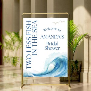 Può includere: Un cartello in metallo dorato con un cartello bianco che recita "TWO LESS FISH IN THE SEA" e "Welcome to AMANDA'S Bridal Shower". Il cartello presenta un design a onda blu ad acquerello e due piccole illustrazioni di uccelli.