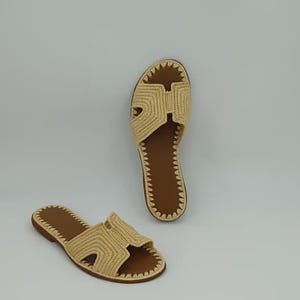 May include: Two beige woven sandals with brown soles are displayed against a white background. The sandals have a distinctive H-shaped design on the top and a stitched border. The open-toe design and neutral colors suggest a casual, summery style.