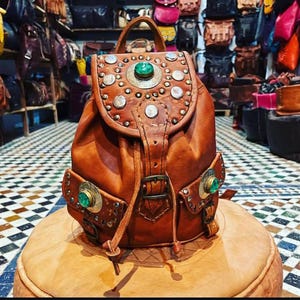 Puede incluir: Mochila de cuero marrón con tachuelas plateadas y una piedra verde central. La bolsa tiene dos bolsillos delanteros con detalles a juego y un asa superior. Descansa sobre una superficie marrón, con otras bolsas en el fondo.