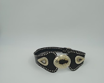 May include: Black leather belt with silver-toned metal accents. The belt features a large oval buckle with a dark stone and decorative silver studs. The belt has a western style.