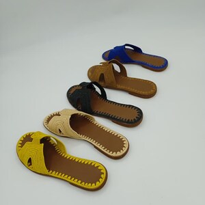 Op de afbeelding: Vijf paar open-teen sandalen in verschillende kleuren: geel, beige, zwart, bruin en blauw. Elke sandaal heeft een geweven bovenwerk en een bruine zool. De sandalen zijn diagonaal geplaatst tegen een witte achtergrond, wat hun ontwerp en kleurvariaties benadrukt.