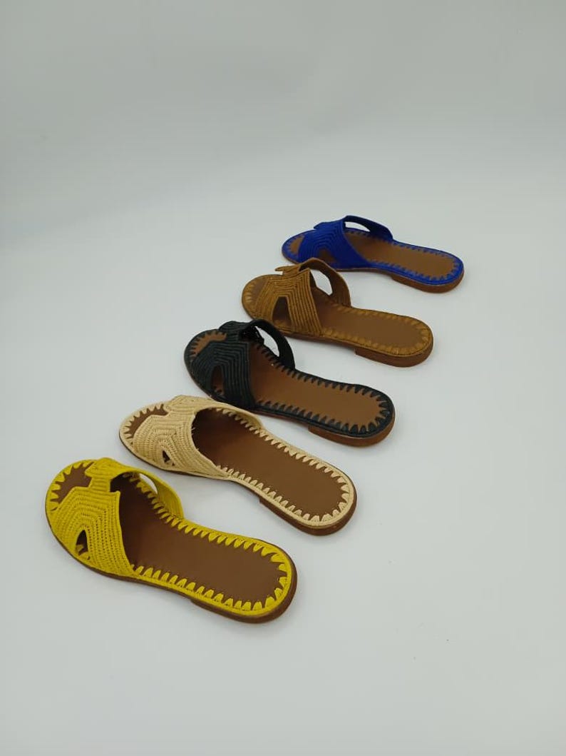 Op de afbeelding: Vijf paar open-teen sandalen in verschillende kleuren: geel, beige, zwart, bruin en blauw. Elke sandaal heeft een geweven bovenwerk en een bruine zool. De sandalen zijn diagonaal geplaatst tegen een witte achtergrond, wat hun ontwerp en kleurvariaties benadrukt.