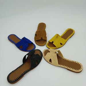 Op de afbeelding: Vijf paar handgemaakte sandalen in verschillende kleuren: blauw, bruin, geel, zwart en beige. Elke sandaal heeft een geweven bovenwerk en een bruine zool met een gestikte rand. De sandalen zijn in een cirkelvormig patroon tegen een witte achtergrond geplaatst.