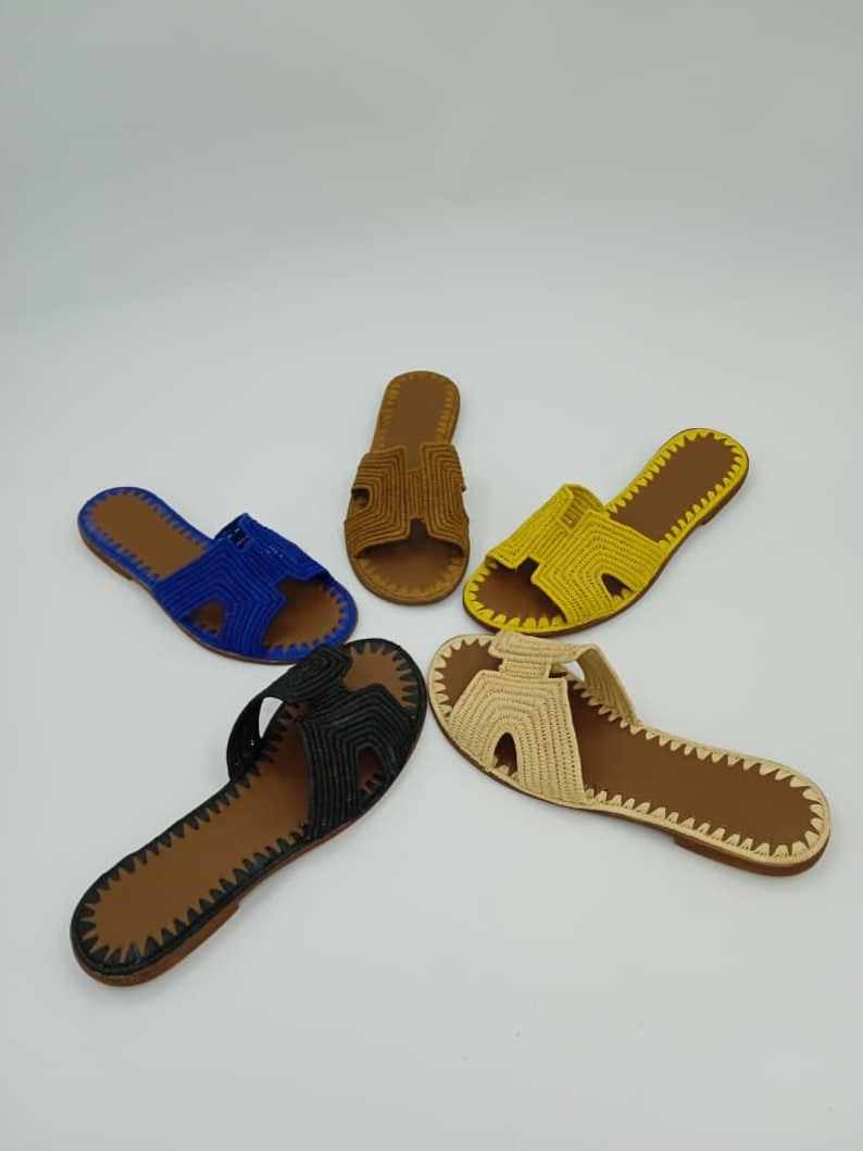 Op de afbeelding: Vijf paar handgemaakte sandalen in verschillende kleuren: blauw, bruin, geel, zwart en beige. Elke sandaal heeft een geweven bovenwerk en een bruine zool met een gestikte rand. De sandalen zijn in een cirkelvormig patroon tegen een witte achtergrond geplaatst.