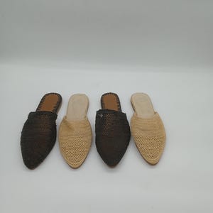 Pode incluir: Quatro mules de ráfia tecida em fila, duas em castanho escuro e duas em bege. Cada sapato tem bico fino e palmilha de couro. Os sapatos são exibidos em fundo branco.