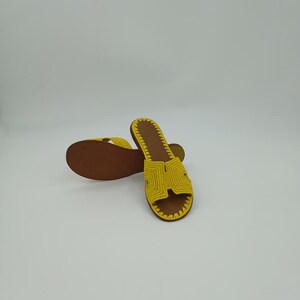 Op de afbeelding: Een paar gele gehaakte sandalen met bruine zolen. De sandalen hebben een eenvoudig ontwerp met een band over de bovenkant van de voet. De zolen zijn donkerbruin van kleur en de sandalen lijken handgemaakt.