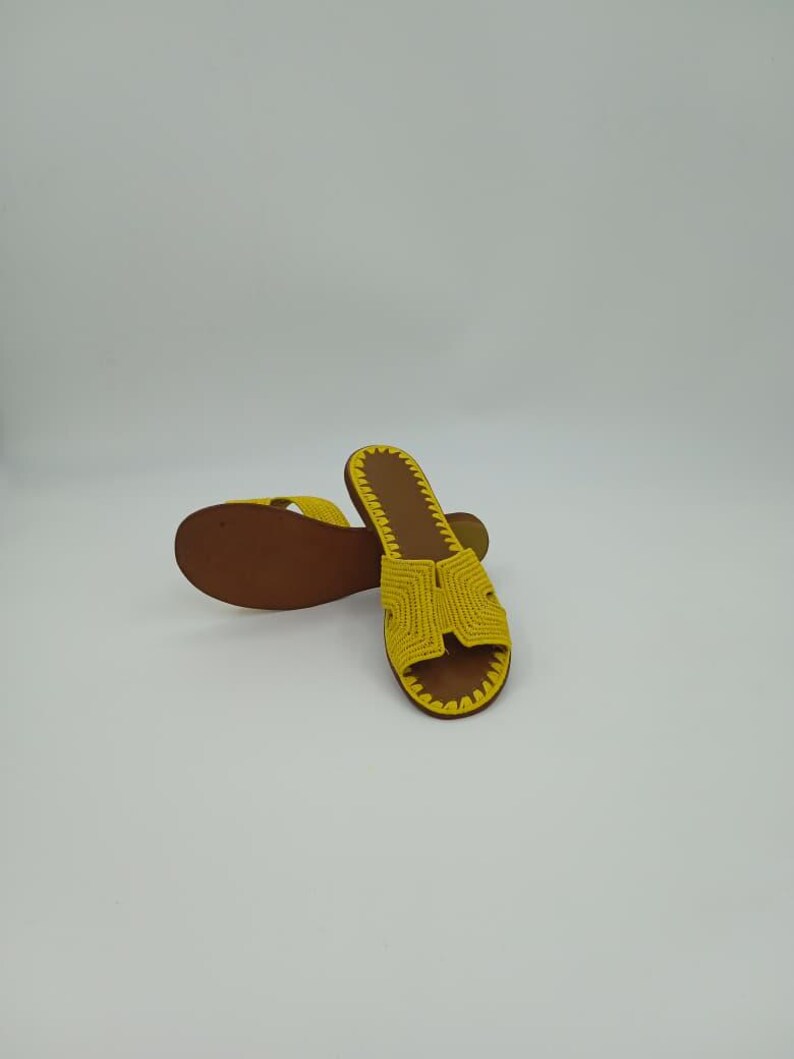 Op de afbeelding: Een paar gele gehaakte sandalen met bruine zolen. De sandalen hebben een eenvoudig ontwerp met een band over de bovenkant van de voet. De zolen zijn donkerbruin van kleur en de sandalen lijken handgemaakt.