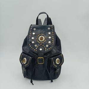 Può includere: Zaino in pelle nera con borchie e abbellimenti argentati e dorati. Lo zaino ha due tasche frontali con chiusure decorative e una chiusura con fibbia. La borsa è dotata di una maniglia superiore e spallacci regolabili.