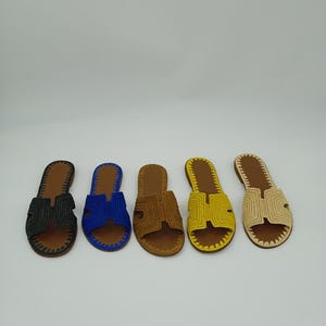 Op de afbeelding: Vijf paar open teensandalen in verschillende kleuren: zwart, blauw, bruin, geel en beige. Elke sandaal heeft een geweven bovenwerk en een bruine leren zool. De sandalen zijn in een rij op een witte achtergrond geplaatst.