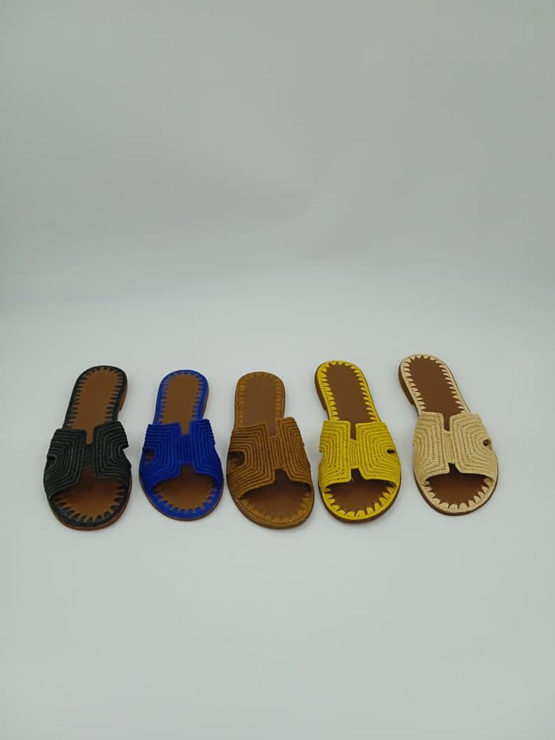 Op de afbeelding: Vijf paar open teensandalen in verschillende kleuren: zwart, blauw, bruin, geel en beige. Elke sandaal heeft een geweven bovenwerk en een bruine leren zool. De sandalen zijn in een rij op een witte achtergrond geplaatst.