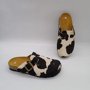 Puede incluir: Dos zuecos con estampado de piel de vaca. Los zuecos tienen un patrón blanco y negro, una suela de corcho y un cierre de hebilla. El interior es de color marrón claro.