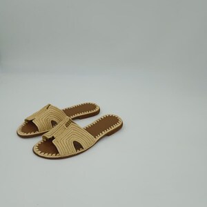 Op de afbeelding: Een paar beige geweven sandalen met bruine zolen. De sandalen hebben een slip-on design met een open teen en een geweven band over de bovenkant. De zolen zijn plat en hebben een gestikte rand. De sandalen worden op een witte achtergrond weergegeven.
