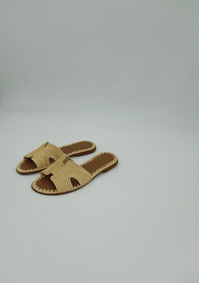 Op de afbeelding: Een paar beige geweven sandalen met bruine zolen. De sandalen hebben een slip-on design met een open teen en een geweven band over de bovenkant. De zolen zijn plat en hebben een gestikte rand. De sandalen worden op een witte achtergrond weergegeven.