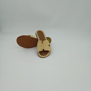 Op de afbeelding: Een paar beige geweven sandalen met een bruine zool. De sandalen hebben een H-vormig ontwerp aan de bovenkant en een gestikte rand. De sandalen worden op een witte achtergrond gepresenteerd.
