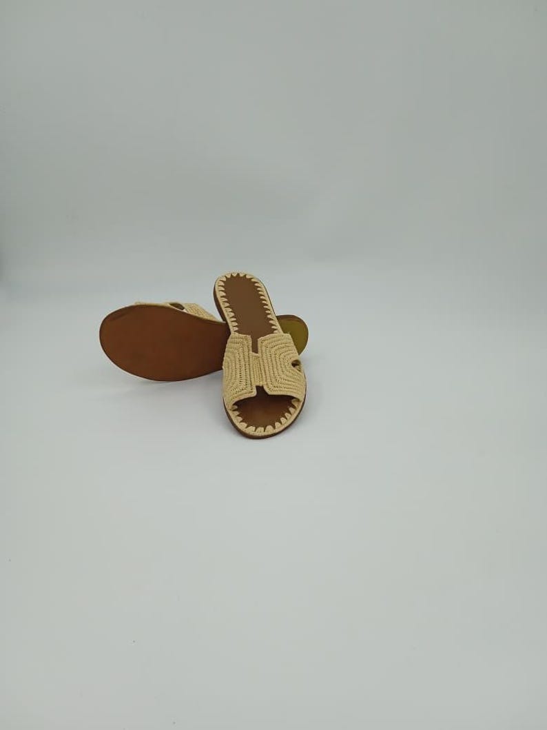 Op de afbeelding: Een paar beige geweven sandalen met een bruine zool. De sandalen hebben een H-vormig ontwerp aan de bovenkant en een gestikte rand. De sandalen worden op een witte achtergrond gepresenteerd.