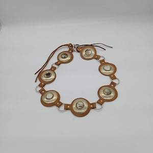 以下が含まれることがあります： シルバーの金属アクセントが付いた茶色の革ベルト。ベルトは、中央にシルバーのディスクと茶色の革の縁取りが付いた円形の装飾要素を備えています。ベルトは、接続されたピースと革のネクタイで設計されています。