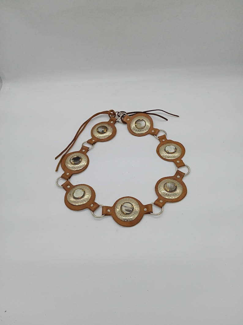 以下が含まれることがあります： シルバーの金属アクセントが付いた茶色の革ベルト。ベルトは、中央にシルバーのディスクと茶色の革の縁取りが付いた円形の装飾要素を備えています。ベルトは、接続されたピースと革のネクタイで設計されています。