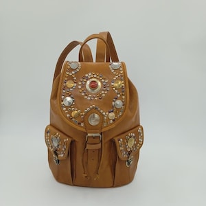 Può includere: Uno zaino in pelle marrone chiaro con borchie metalliche decorative e una pietra rossa centrale. Lo zaino ha due tasche frontali, spallacci regolabili e una chiusura con coulisse. La borsa è progettata con un'estetica vintage.