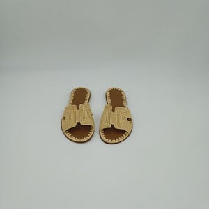 Op de afbeelding: Een paar beige geweven sandalen met bruine binnenzolen. De sandalen hebben een H-vormig ontwerp over de bovenkant en een gestikte rand. Het schoeisel wordt op een witte achtergrond weergegeven.