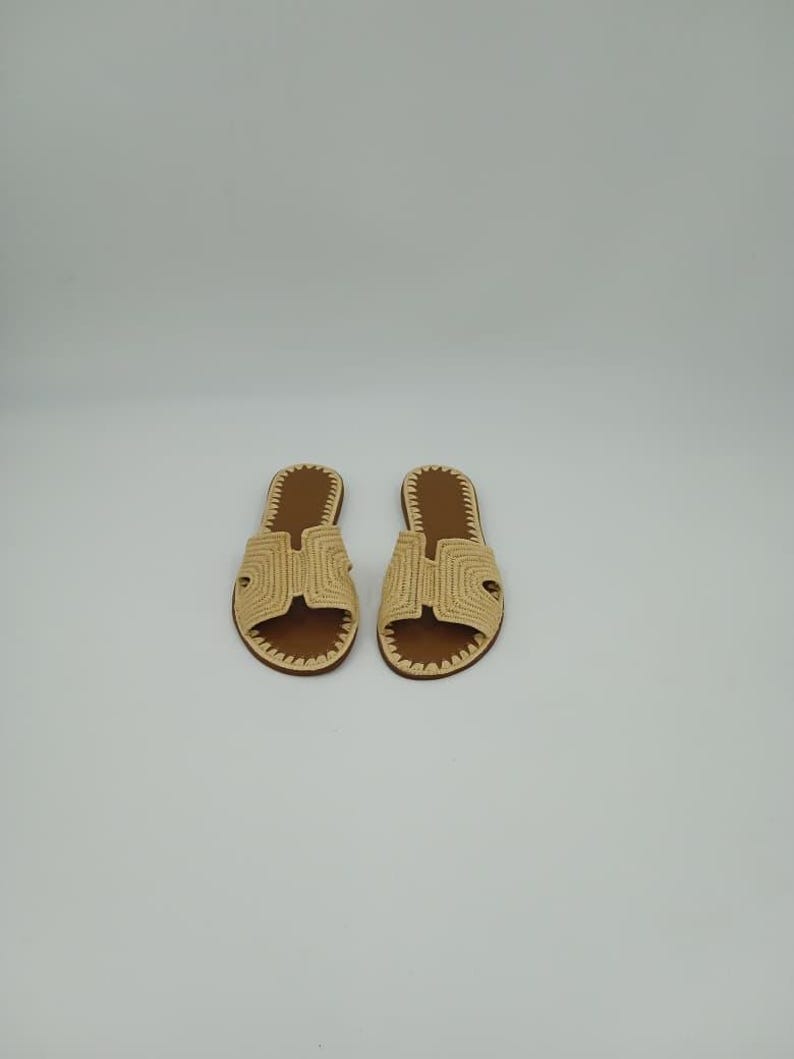 Op de afbeelding: Een paar beige geweven sandalen met bruine binnenzolen. De sandalen hebben een H-vormig ontwerp over de bovenkant en een gestikte rand. Het schoeisel wordt op een witte achtergrond weergegeven.