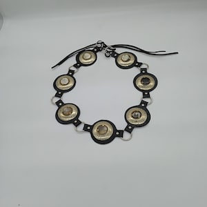 以下が含まれることがあります： シルバーカラーの金属アクセントと円形装飾が特徴の黒いベルト。各円形装飾には中央に石があります。ベルトは黒い革ストラップで接続されており、白い背景に表示されています。