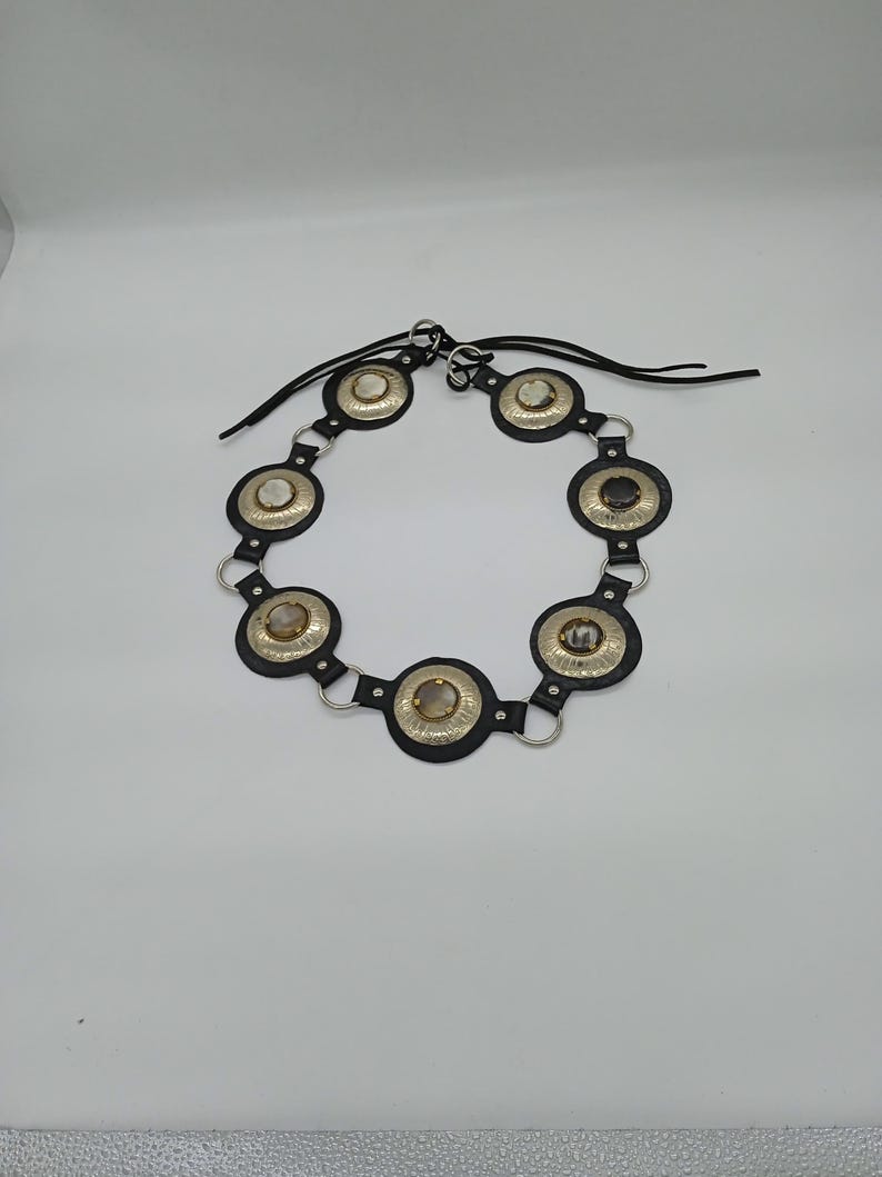 以下が含まれることがあります： シルバーカラーの金属アクセントと円形装飾が特徴の黒いベルト。各円形装飾には中央に石があります。ベルトは黒い革ストラップで接続されており、白い背景に表示されています。