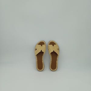 Op de afbeelding: Een paar beige geweven sandalen met bruine zolen. De sandalen hebben een eenvoudig ontwerp met een band over de bovenkant en een textuurpatroon. De zolen zijn donkerbruin.