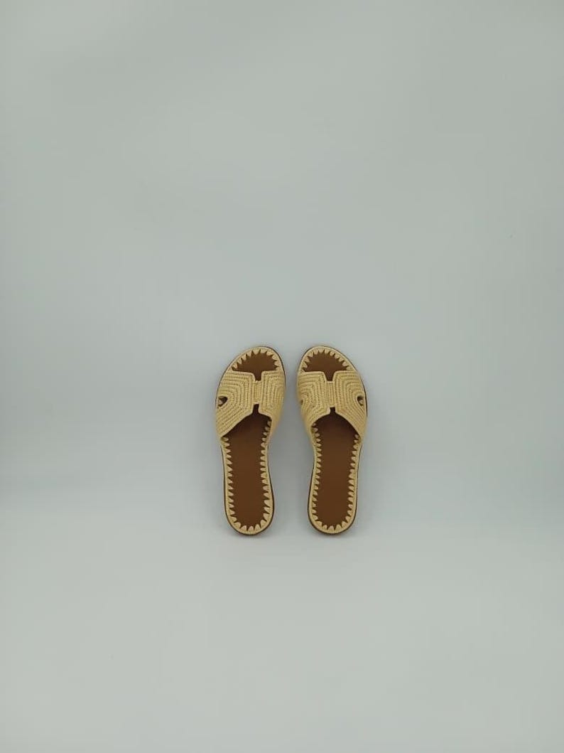 Op de afbeelding: Een paar beige geweven sandalen met bruine zolen. De sandalen hebben een eenvoudig ontwerp met een band over de bovenkant en een textuurpatroon. De zolen zijn donkerbruin.