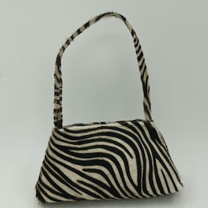Peut inclure: Un petit sac à main doté d'un motif zébré texturé en noir et blanc. Le sac présente une forme trapézoïdale structurée avec une bandoulière unique assortie au motif zébré, fixée sur le dessus pour un design animalier cohérent.