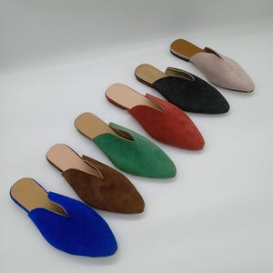 Puede incluir: Una colección de zapatos sin cordones de ante en varios colores, incluyendo azul, marrón, verde, rojo, negro y rosa. Cada zapato presenta una puntera y una plantilla de cuero marrón en contraste. Los zapatos están colocados sobre una superficie blanca.