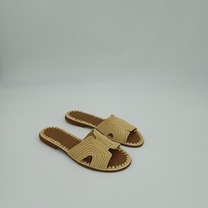 Op de afbeelding: Een paar beige geweven sandalen met bruine zolen. De sandalen hebben een open teenontwerp en een geweven band over de bovenkant. De zolen zijn donkerbruin, tegen een witte achtergrond.