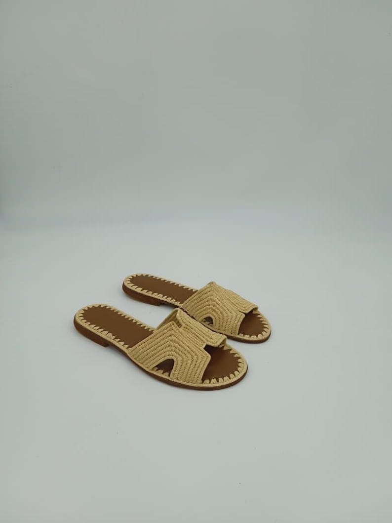 Op de afbeelding: Een paar beige geweven sandalen met bruine zolen. De sandalen hebben een open teenontwerp en een geweven band over de bovenkant. De zolen zijn donkerbruin, tegen een witte achtergrond.