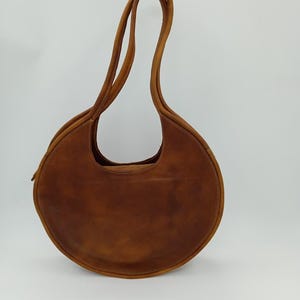 May include: A brown leather handbag with a circular design. The bag has a rounded shape with a long, curved handle. The leather has visible grain and stitching along the edges. The bag is set against a white background.