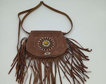 Può includere: Borsa a tracolla in pelle marrone con chiusura a patta e lunghe frange. La borsa presenta un design circolare decorativo con una pietra gialla e piccoli accenti bianchi. Il cinturino è anch'esso in pelle marrone.