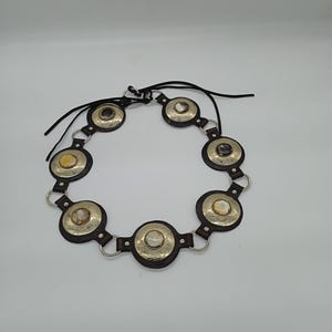 以下が含まれることがあります： シルバーカラーの金属アクセントが付いた、濃い茶色の革製ベルト。ベルトは、中央に石または貝殻のような象嵌が施された円形の装飾要素が特徴です。ベルトはシルバーリングと黒革ストラップで接続されています。
