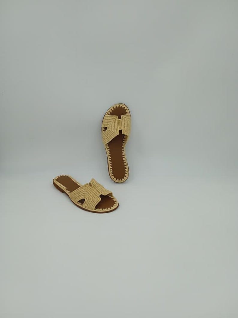 May include: Two beige woven sandals with brown soles are displayed against a white background. The sandals have a distinctive H-shaped design on the top and a stitched border. The open-toe design and neutral colors suggest a casual, summery style.
