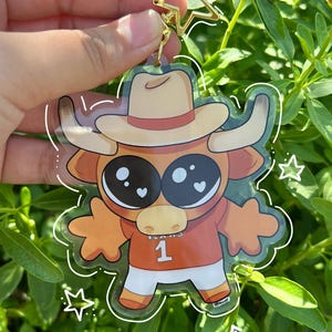 May include: A clear acrylic keychain featuring a cartoon longhorn wearing a cowboy hat and jersey with the number 1. The keychain is 3.5 inches in size and has the text "HOOK'EM KEYCHAIN". A gold star-shaped charm is attached.
