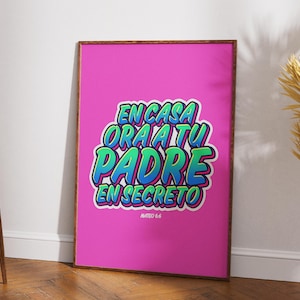 May include: A framed poster with the text "EN CASA ORA A TU PADRE EN SECRETO" in a colorful, bubble-style font on a bright pink background. The text is outlined in blue and green. The poster is leaning against a white wall.