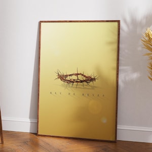 Op de afbeelding: Een ingelijste print met een doornenkroon tegen een gele achtergrond. De woorden "REY DE REYES" staan eronder. De print hangt aan een muur naast een houten stoel en gedroogde planten.